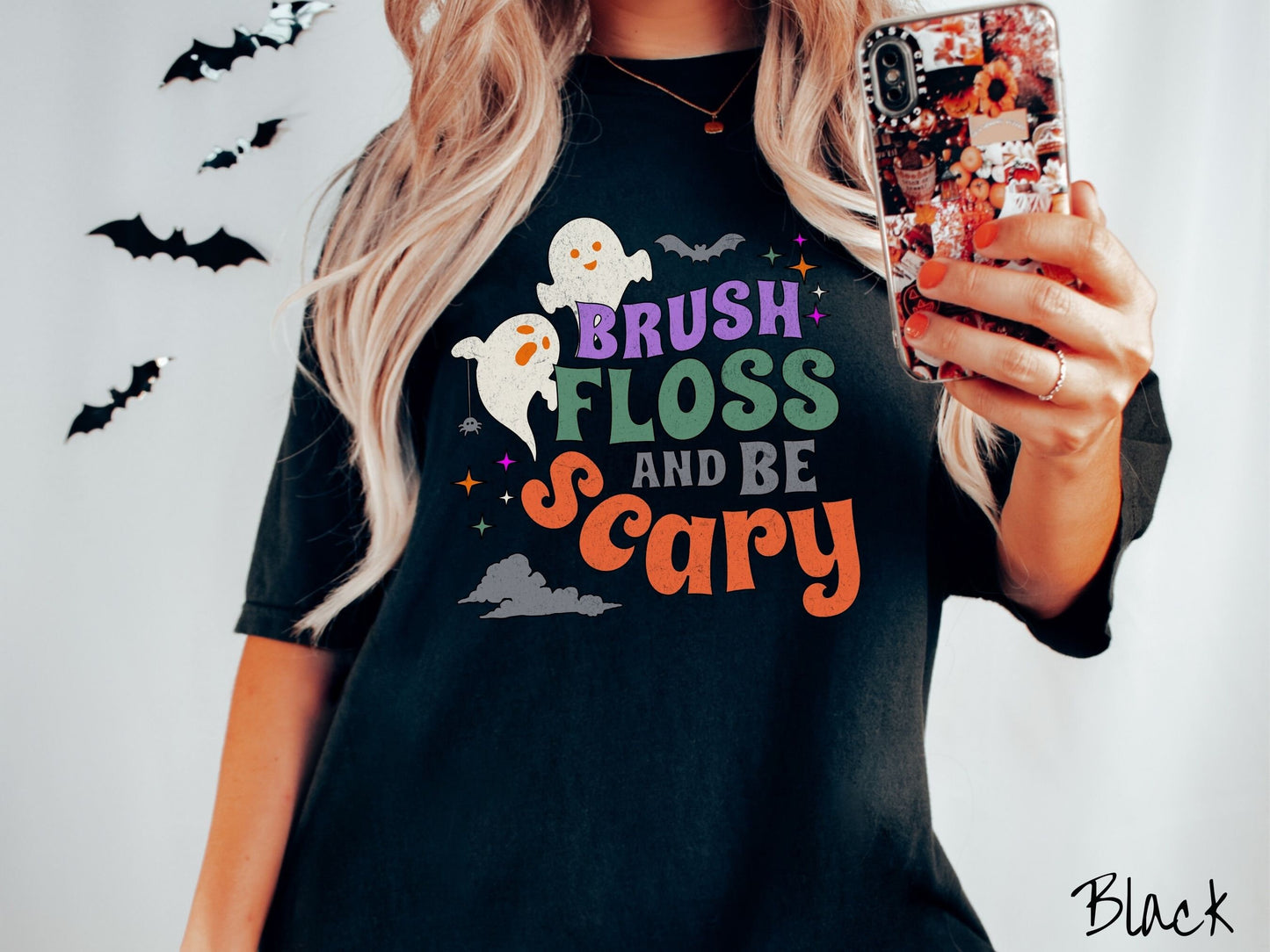 A woman wearing a cute, vintage black colored Comfort Colors T-shirt with the text Brush Floss and Be Scary in colorful font. There are orange-eyed white ghosts flying around and black bats and twinkling stars in the sky.