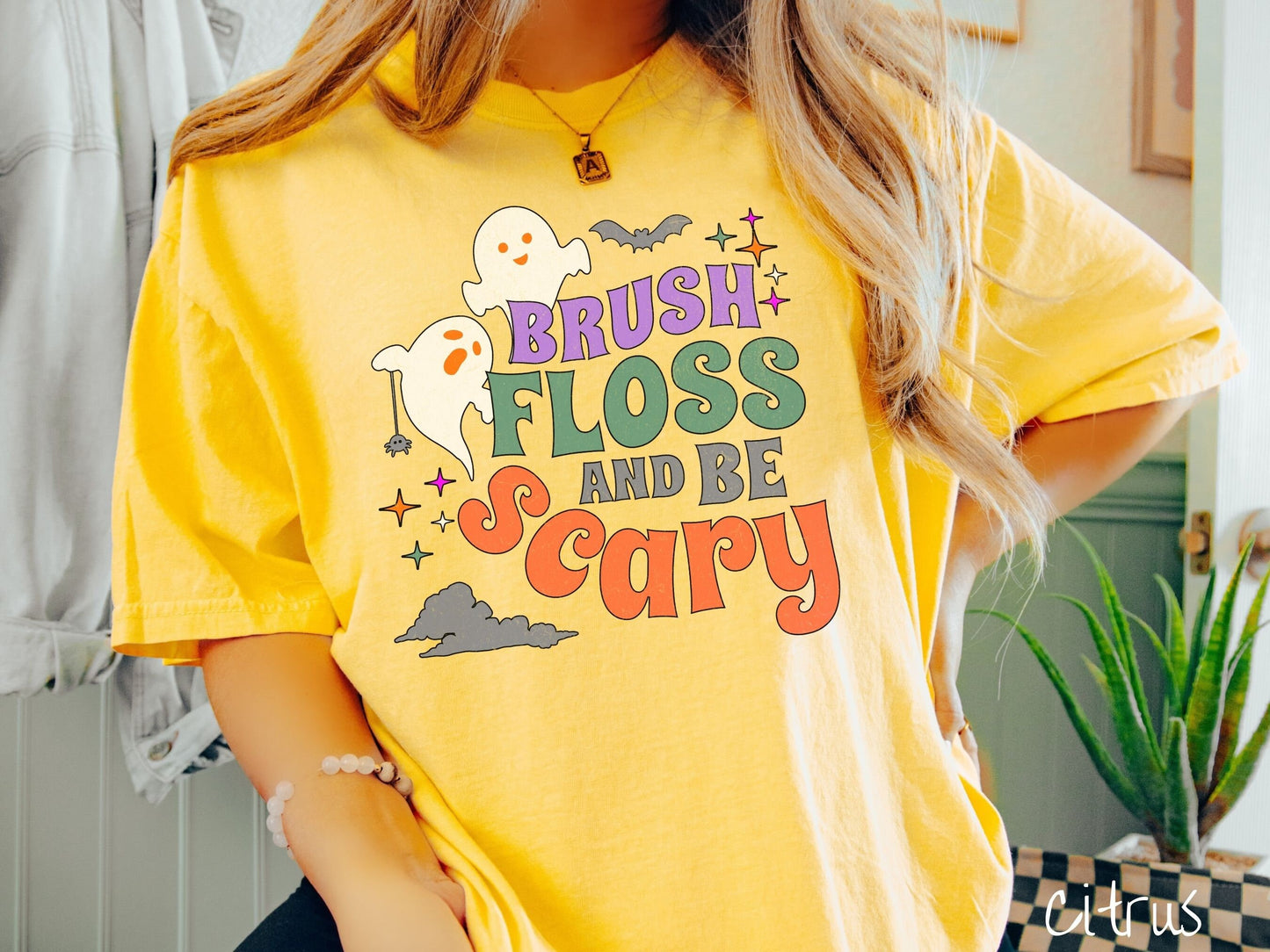 A woman wearing a cute, vintage citrus colored Comfort Colors T-shirt with the text Brush Floss and Be Scary in colorful font. There are orange-eyed white ghosts flying around and black bats and twinkling stars in the sky.