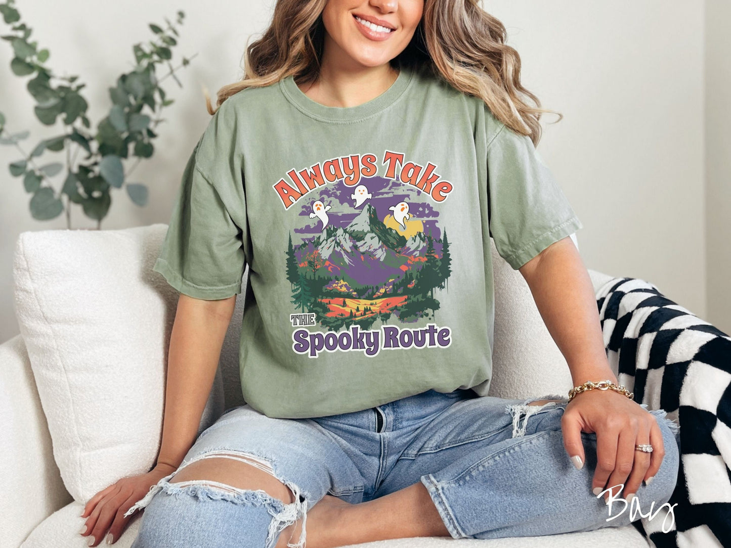 A woman wearing a cute, vintage bay colored Comfort Colors T-shirt with the text Always Take the Spooky Route in colorful font. There are three white ghosts with orange faces flying above a mountain landscape with rolling hills and green forest.
