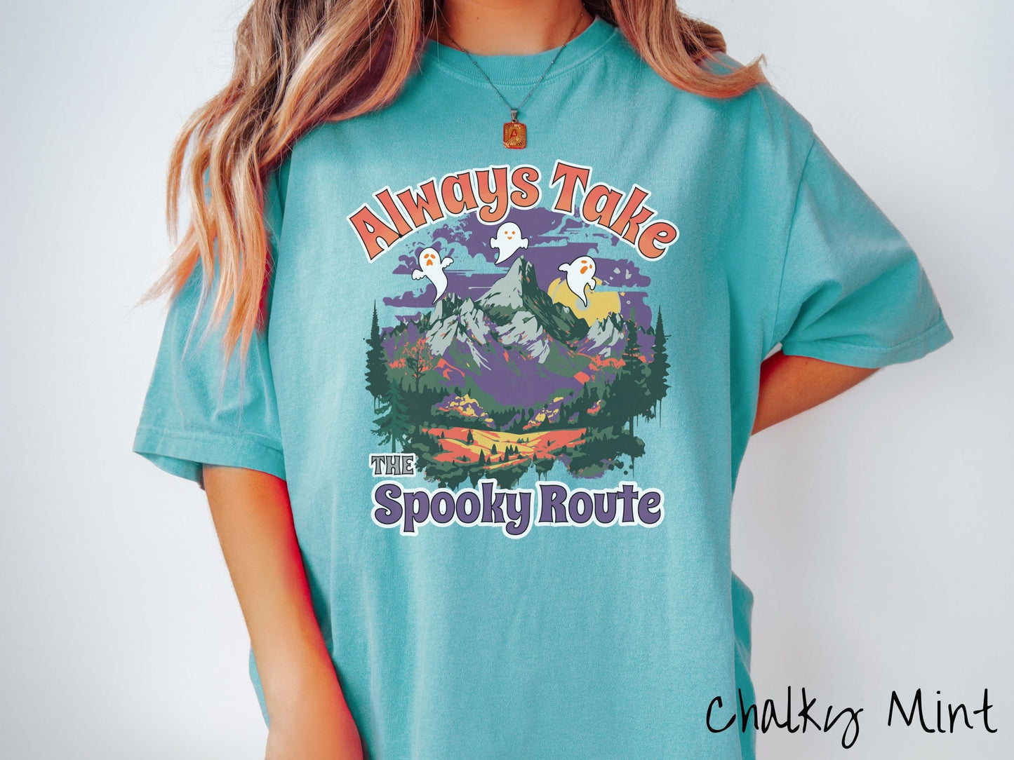 A woman wearing a cute, vintage chalky mint colored Comfort Colors T-shirt with text Always Take the Spooky Route in colorful font. There are three white ghosts with orange faces flying above a mountain landscape with rolling hills and green forest.