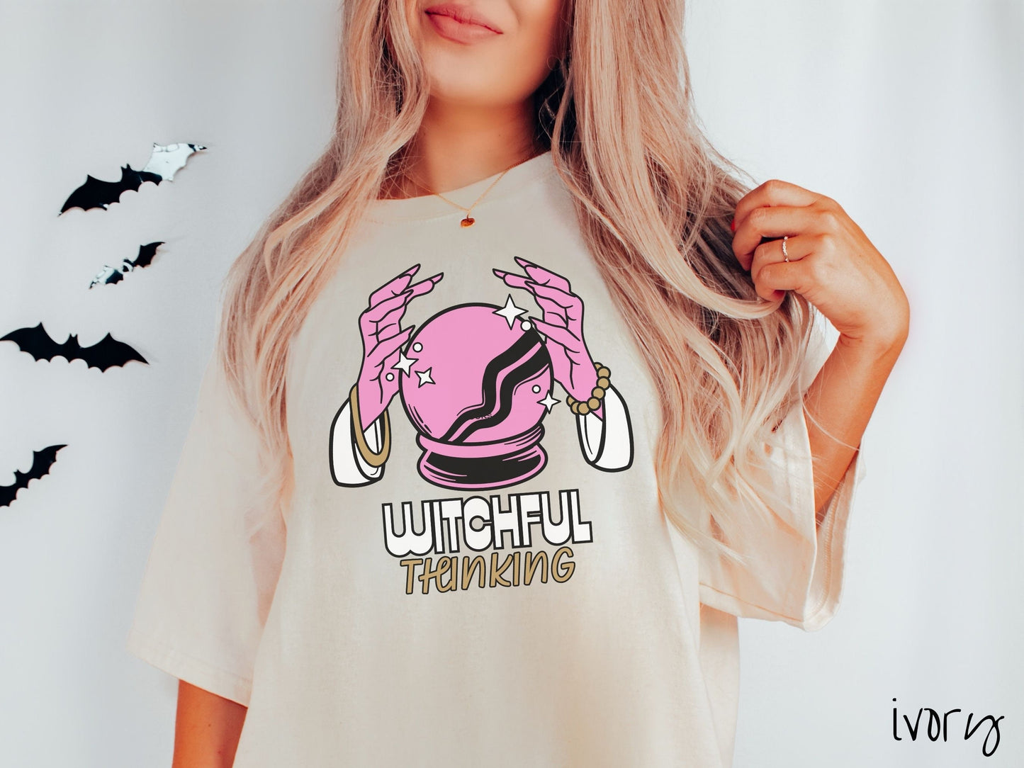 A woman wearing a cute, vintage ivory colored Comfort Colors T-shirt with the text Witchful Thinking in white and gold font. Above is a pink crystal ball, and there are two pink witch hands with jewelry hovering around the crystall ball.