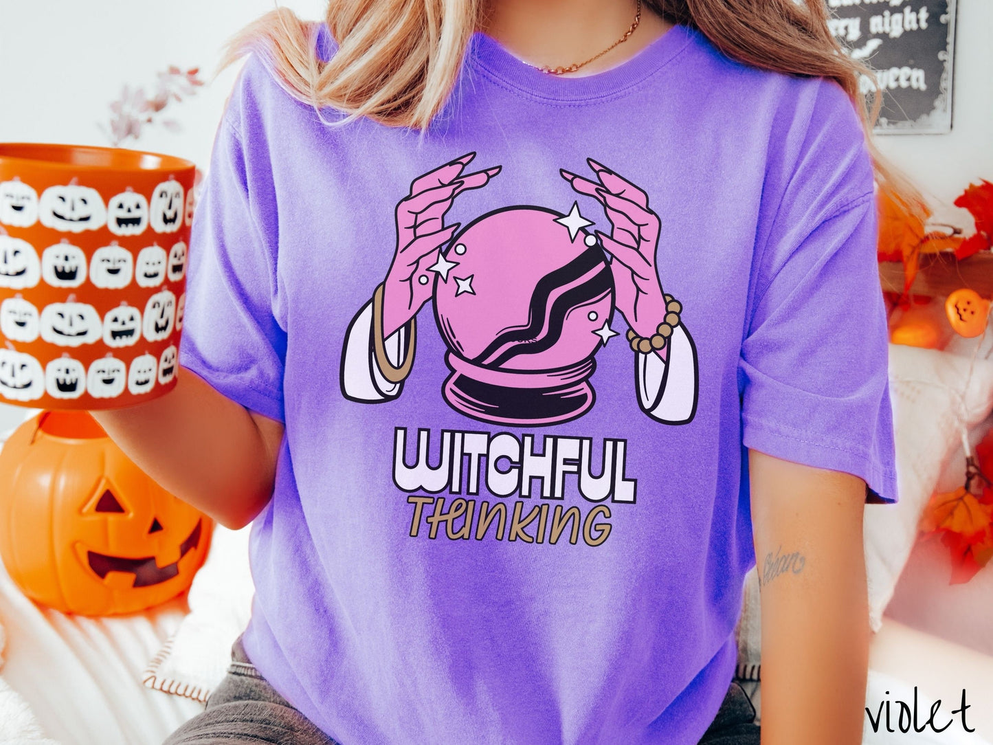 A woman wearing a cute, vintage violet colored Comfort Colors T-shirt with the text Witchful Thinking in white and gold font. Above is a pink crystal ball, and there are two pink witch hands with jewelry hovering around the crystall ball.