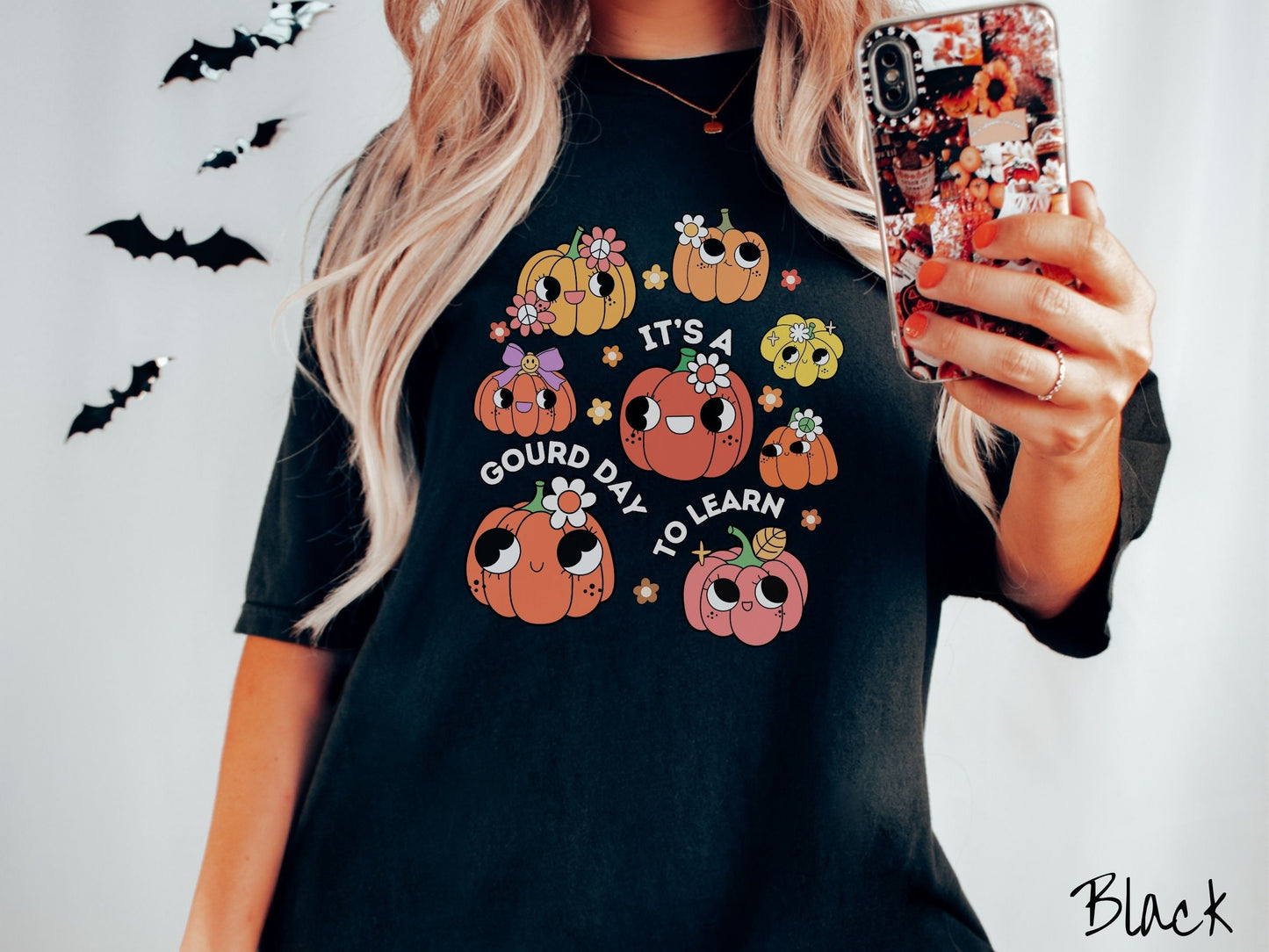 A woman wearing a cute, vintage black colored Comfort Colors T-shirt with the text Its a Gourd Day to Learn in white font. There is a three by three grid of orange, yellow, and pink pumpkins with girly eyes, wearing flowers and bows.