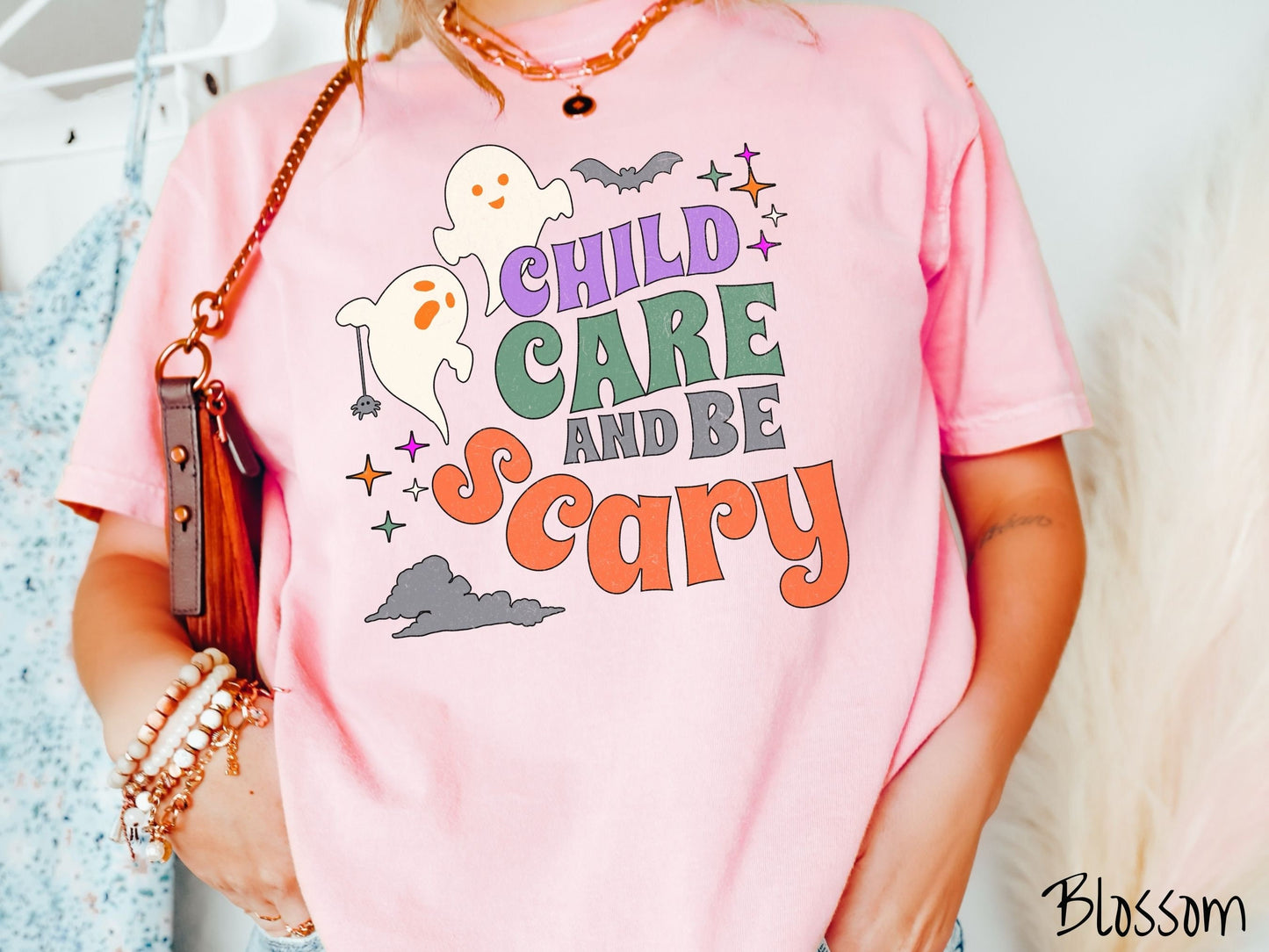 A woman wearing a cute, vintage blossom colored Comfort Colors T-shirt with the text Child Care and Be Scary in colorful font. There are orange-eyed white ghosts flying around and black bats and twinkling stars in the sky.