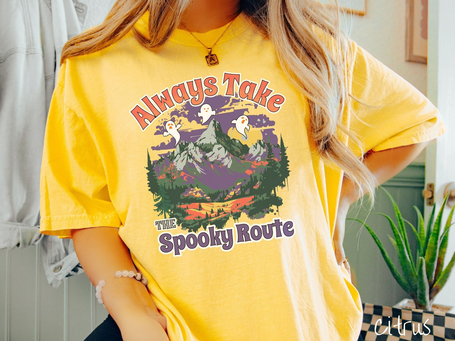 A woman wearing a cute, vintage citrus colored Comfort Colors T-shirt with the text Always Take the Spooky Route in colorful font. There are three white ghosts with orange faces flying above a mountain landscape with rolling hills and green forest.