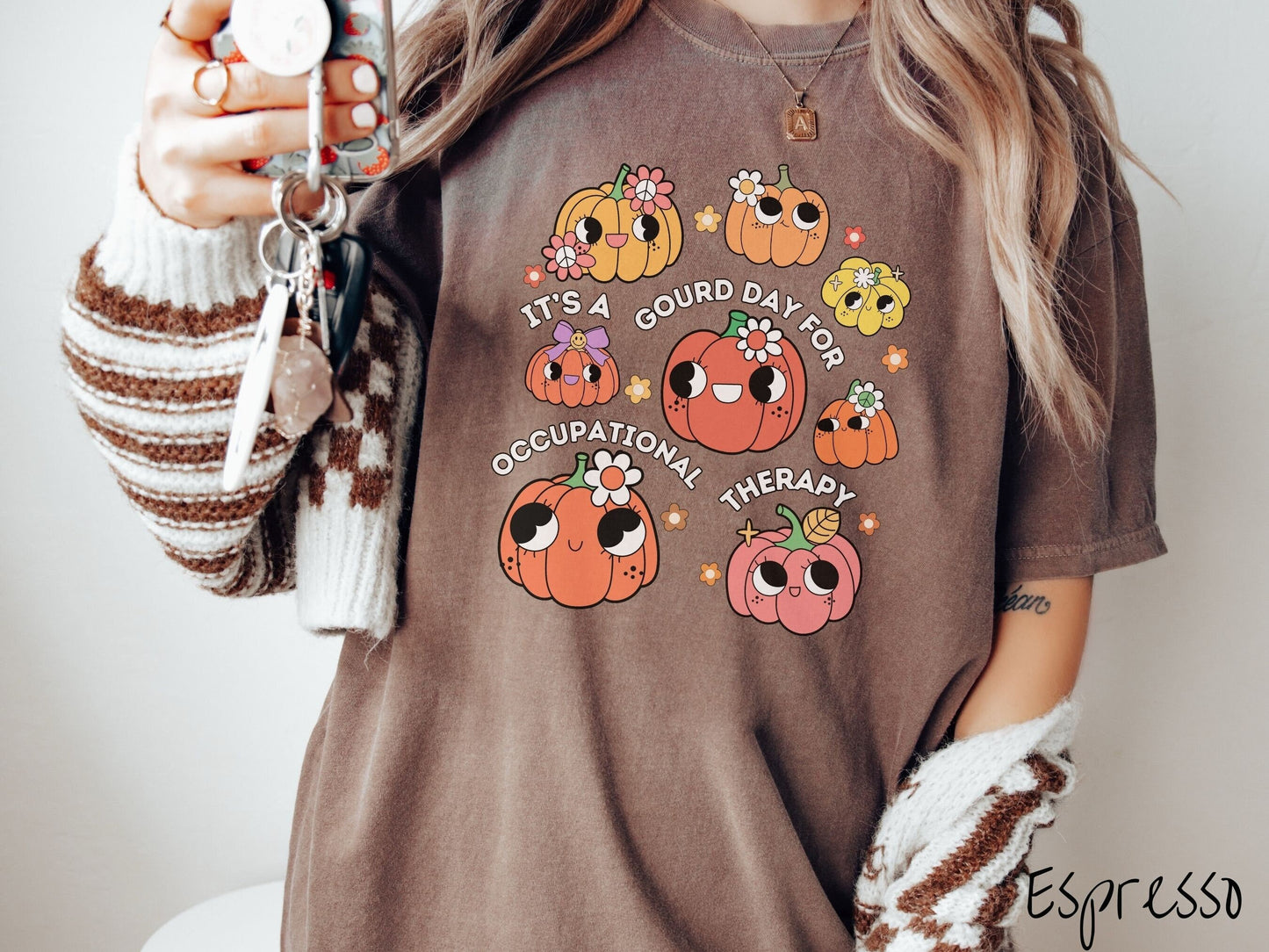 A woman wearing a cute, vintage espresso colored Comfort Colors T-shirt with text Its a Gourd Day for Occupational Therapy in white font. There is a three by three grid of orange, yellow, and pink pumpkins with girly eyes, wearing flowers and bows.