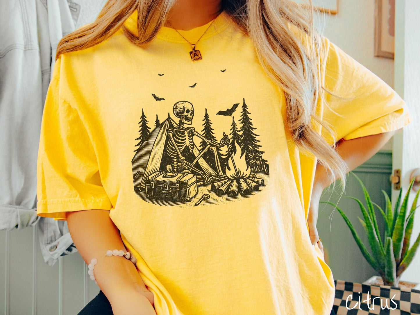 A woman wearing a cute, vintage citrus colored Comfort Colors T-shirt with a skeleton sitting in a tent building a fire next to a cooler out in the woods. The campsite is surrounded by evergreen trees and there are bats flying above.