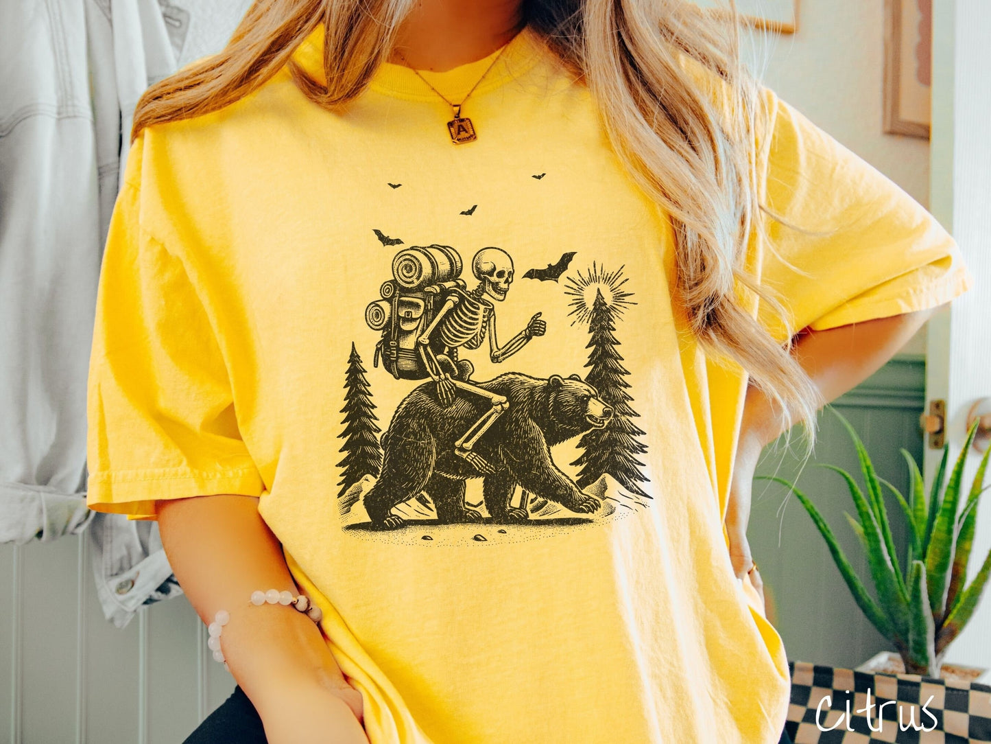 A woman wearing a cute, vintage citrus colored Comfort Colors T-shirt with a smiling skeleton wearing a camping backpack and riding a bear in the woods. There are trees in the background in front of the moon and black bats in the air.