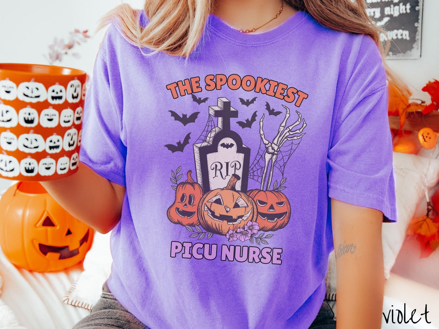 A woman wearing a cute, vintage violet colored Comfort Colors T-shirt with text The Spookiest PICU Nurse. There are three smiling carved pumpkins sitting in front of a tombstone with a skeleton arm coming out of the ground and black bats in the air.