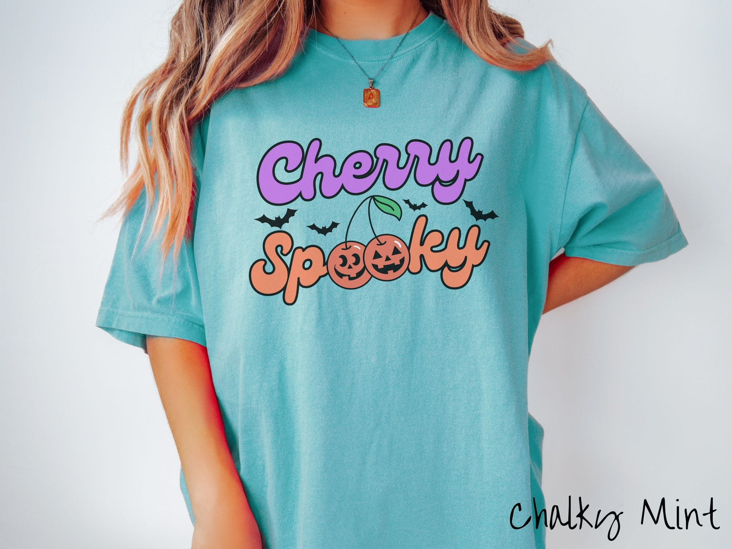 A woman wearing a vintage, chalky mint colored Comfort Colors t-shirt with the text Cherry Spooky in purple and orange font. The two letter O’s in Spooky are smiling jack o'lantern-themed cherries, and there are black bats flying in the background.