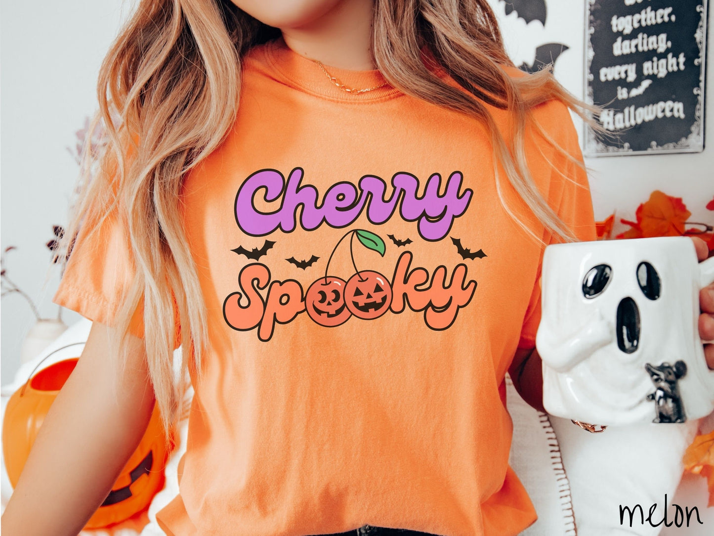A woman wearing a vintage, melon colored Comfort Colors t-shirt with the text Cherry Spooky in purple and orange font. The two letter O’s in Spooky are smiling jack o'lantern-themed cherries, and there are black bats flying in the background.