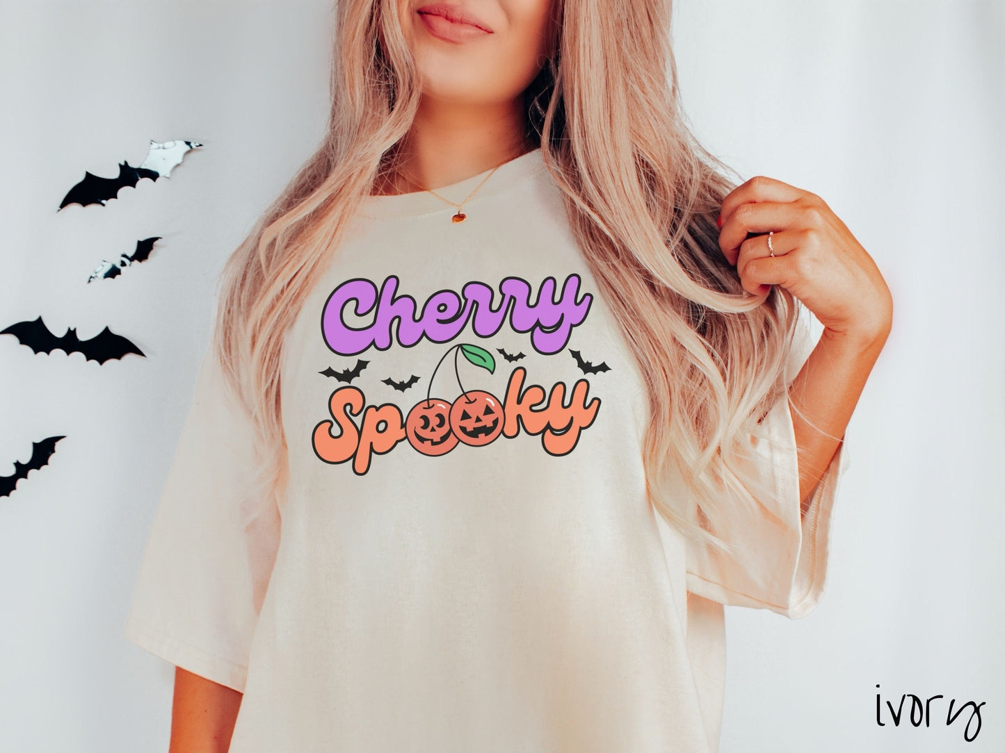A woman wearing a vintage, ivory colored Comfort Colors t-shirt with the text Cherry Spooky in purple and orange font. The two letter O’s in Spooky are smiling jack o'lantern-themed cherries, and there are black bats flying in the background.