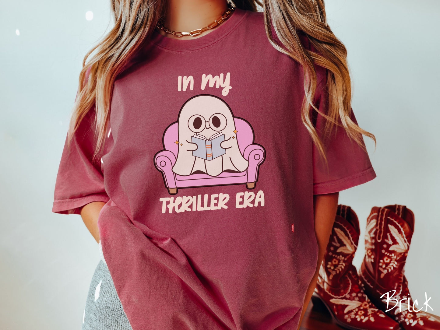 A woman wearing a vintage, brick colored Comfort Colors t-shirt with the text In My Thriller Era across the front in ivory font. In between the text is a young ghost with glasses reading a book and sitting in a pink chair.