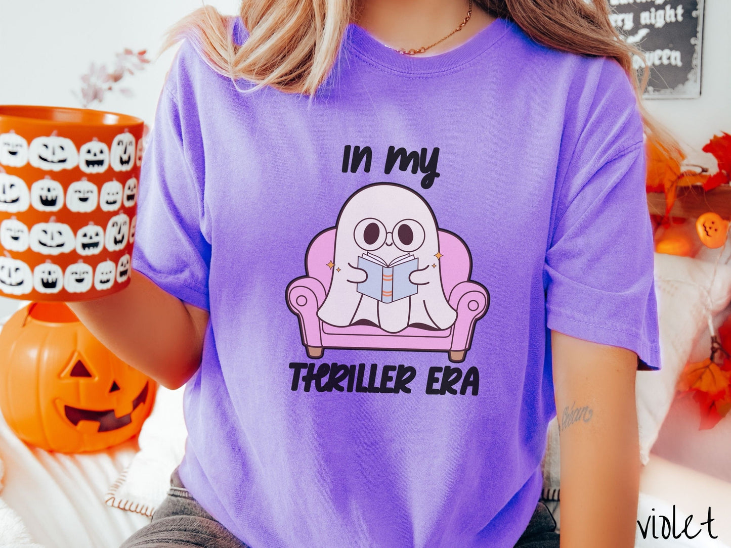 A woman wearing a vintage, violet colored Comfort Colors t-shirt with the text In My Thriller Era across the front in black font. In between the text is a young ghost with glasses reading a book and sitting in a pink chair.