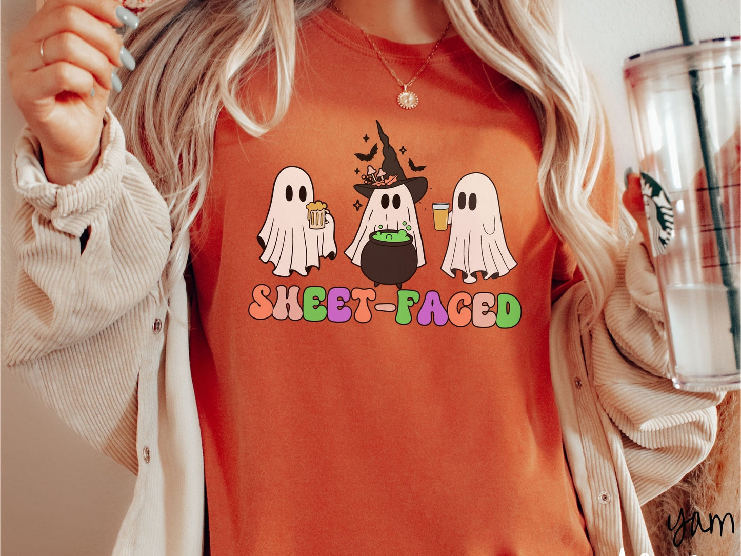 A woman wearing a vintage, yam colored Comfort Colors t-shirt with the text Sheet-Faced in colorful font across the front. Above are three white ghosts drinking beers and standing around a black cauldron with a bubbling green liquid in it.