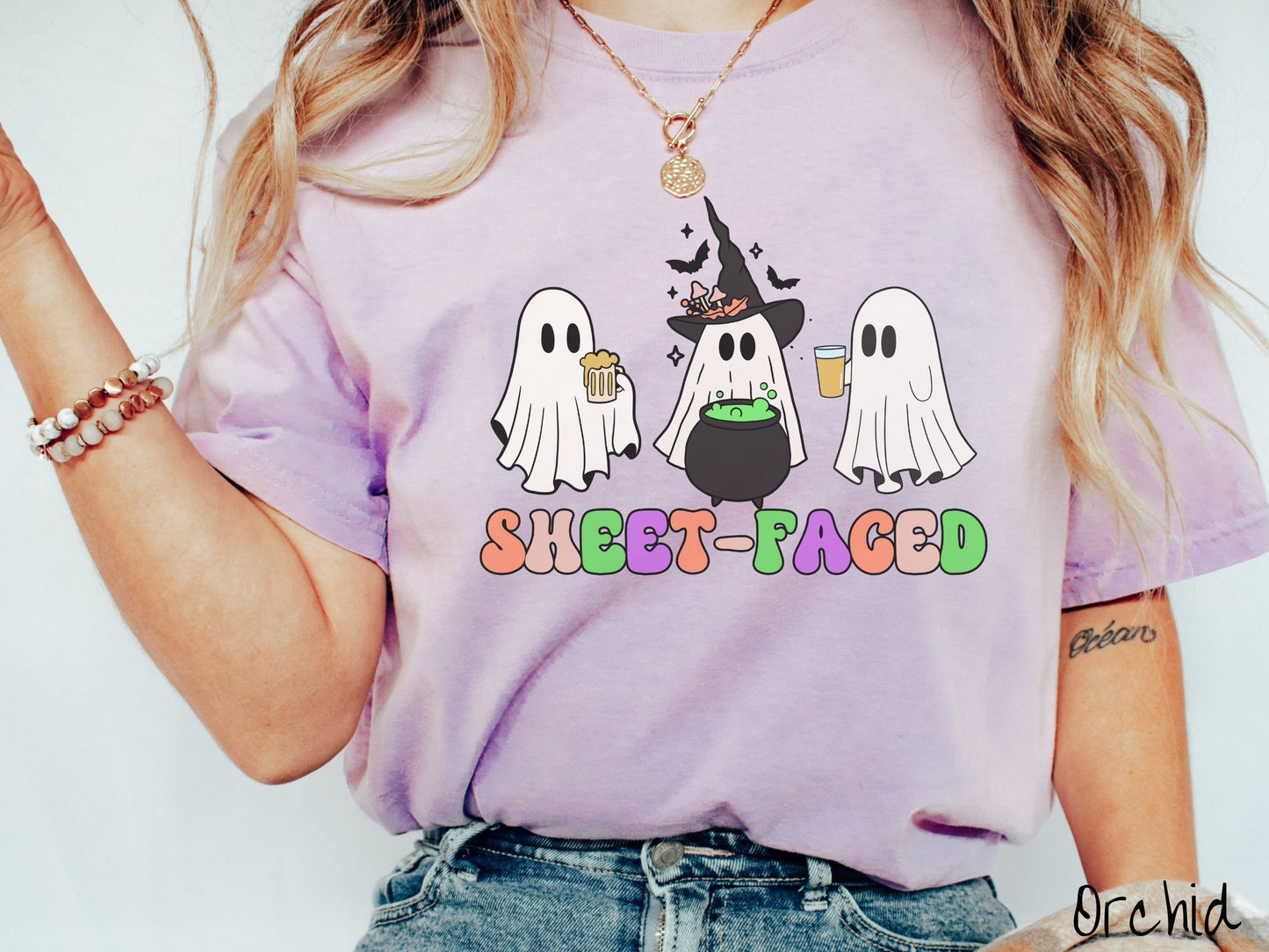 A woman wearing a vintage, orchid colored Comfort Colors t-shirt with the text Sheet-Faced in colorful font across the front. Above are three white ghosts drinking beers and standing around a black cauldron with a bubbling green liquid in it.