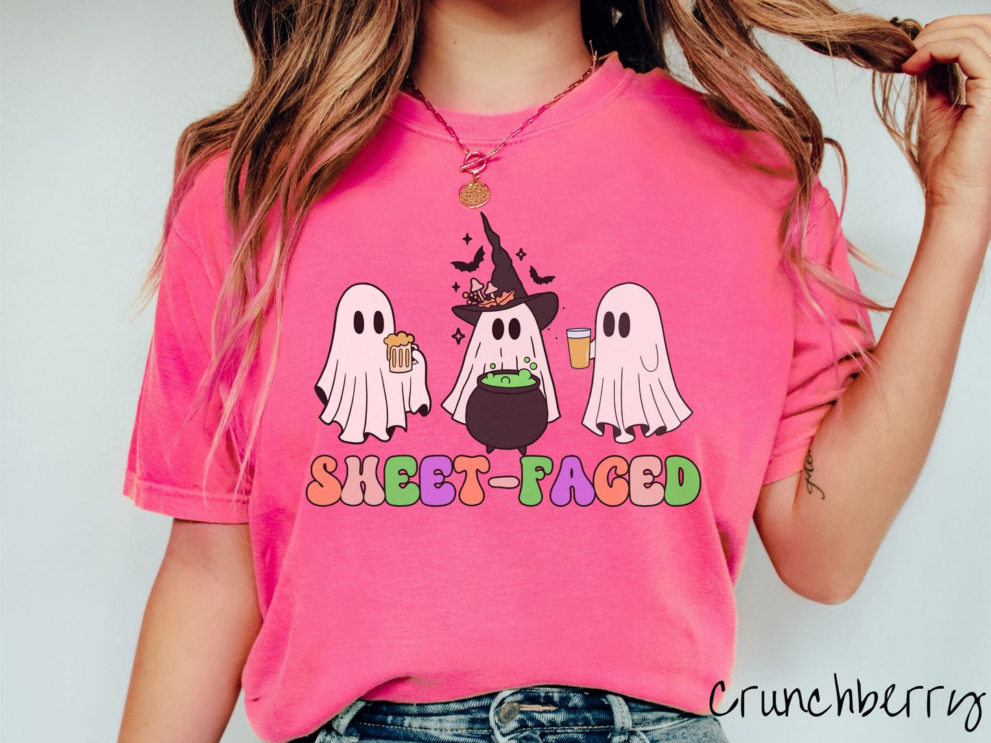 A woman wearing a vintage, crunchberry colored Comfort Colors t-shirt with the text Sheet-Faced in colorful font across the front. Above are three white ghosts drinking beers and standing around a black cauldron with a bubbling green liquid in it.