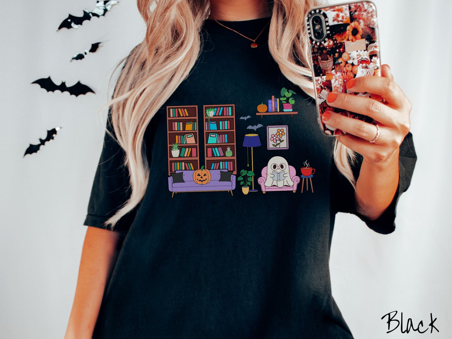 A woman wearing a vintage, black colored Comfort Colors t-shirt with a cozy Halloween-themed room. A young white ghost is reading a book in a chair next to a coffee and colorful bookcase.. An orange pumpkin sits on the couch and bats are in the air.