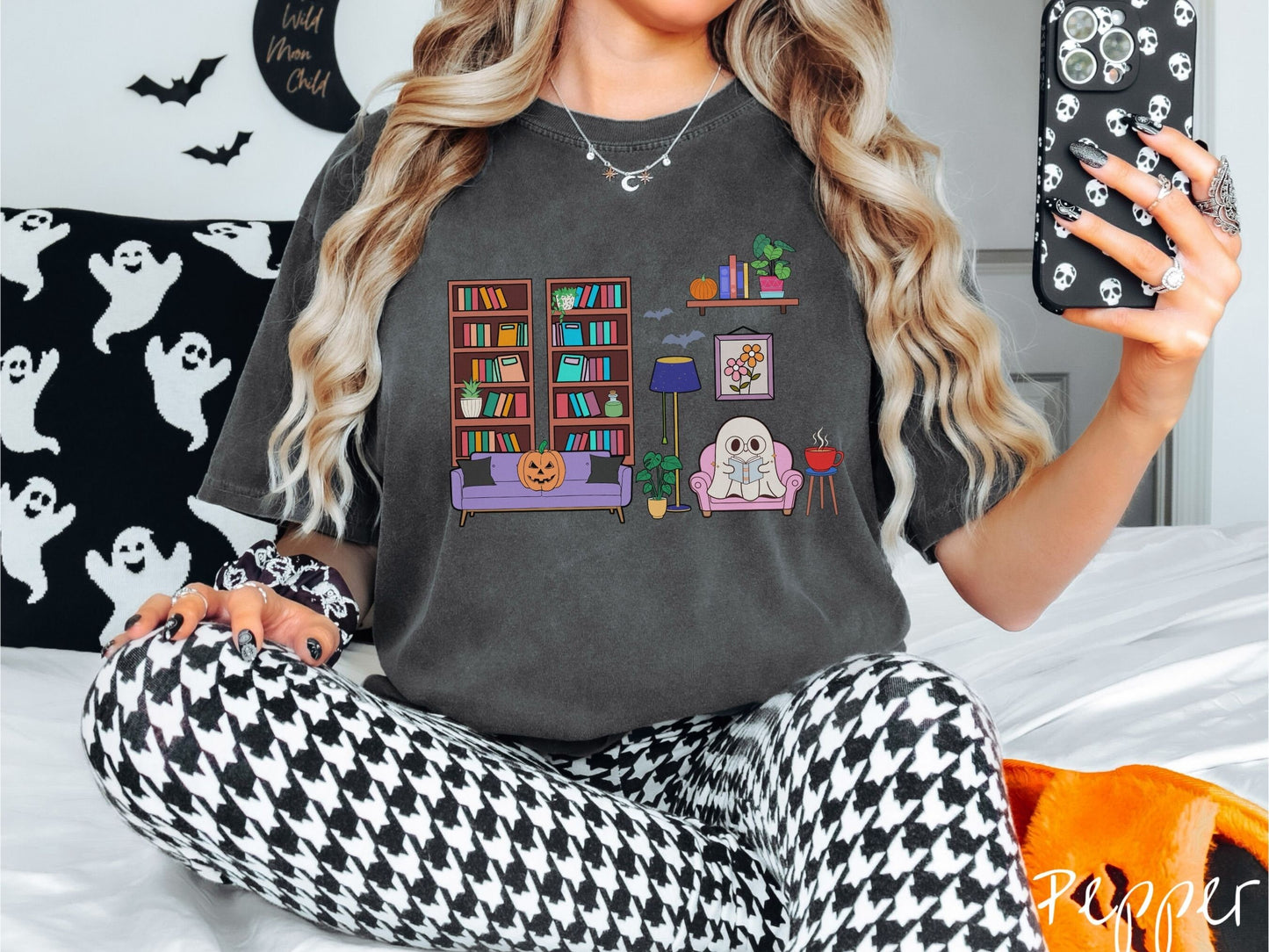 A woman wearing a vintage, pepper colored Comfort Colors t-shirt with a cozy Halloween-themed room. A young white ghost is reading a book in a chair next to a coffee and colorful bookcase.. An orange pumpkin sits on the couch and bats are in the air.