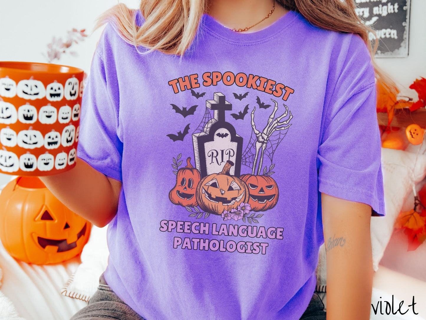 A woman wearing a cute, vintage violet colored Comfort Colors T-shirt with text The Spookiest Speech Language Pathologist. There are three pumpkins sitting in front of a tombstone with a skeleton arm coming out of the ground and bats in the air.