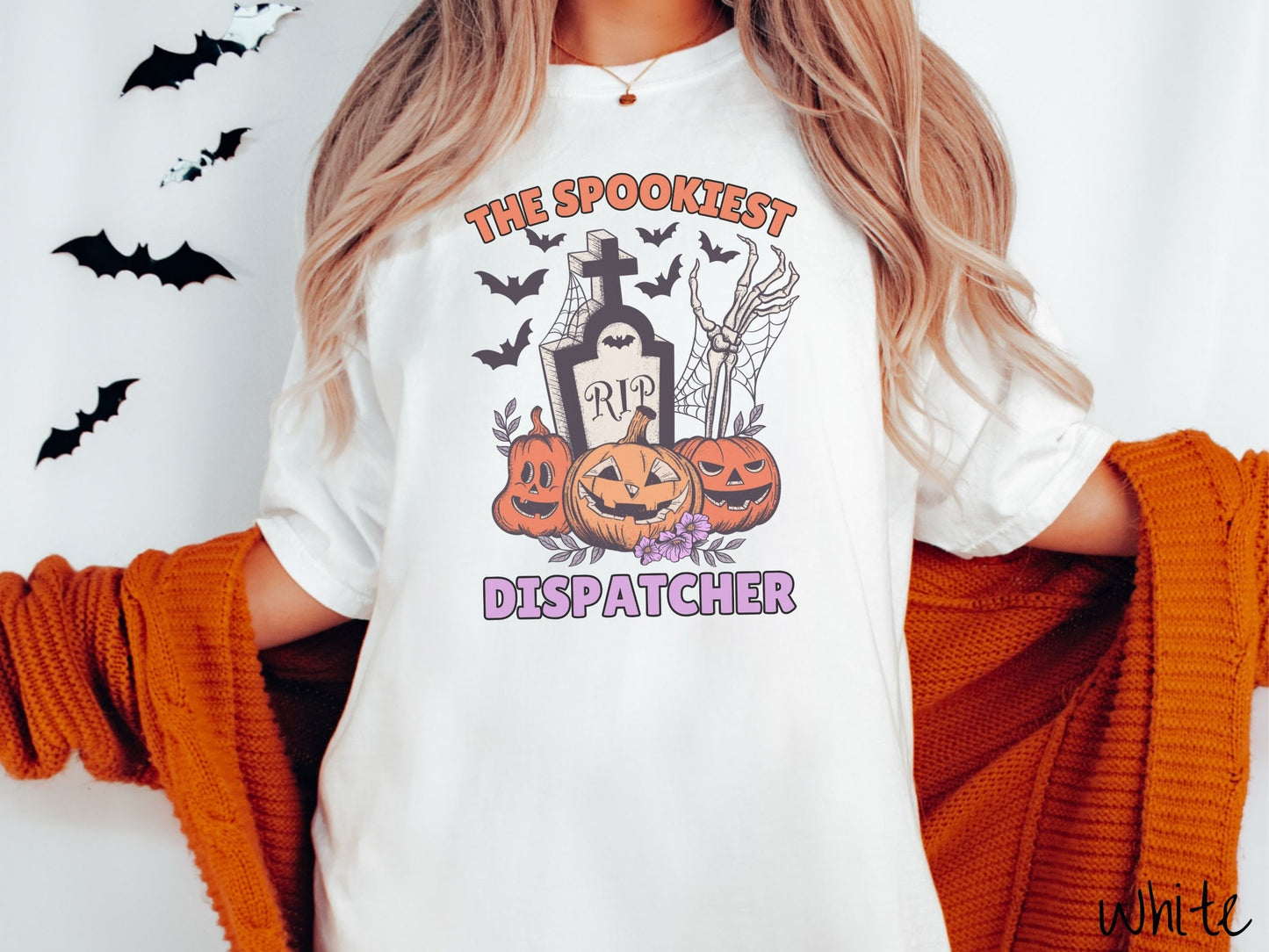 A woman wearing a cute, vintage white colored Comfort Colors T-shirt with text The Spookiest Dispatcher. There are three smiling carved pumpkins sitting in front of a tombstone with a skeleton arm coming out of the ground and black bats in the air.