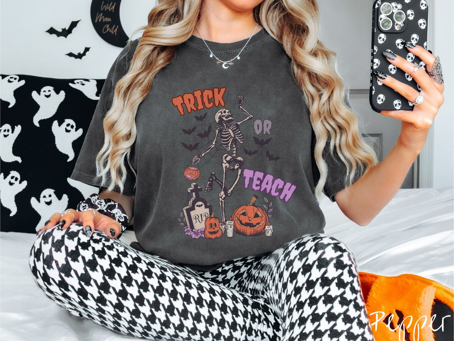A woman wearing a cute, vintage pepper colored Comfort Colors T-shirt with text Trick or Teach in purple and orange font. There is a skeleton holding a pumpkin candy bucket dancing among spider webs and black bats over a tombstone and pumpkins.