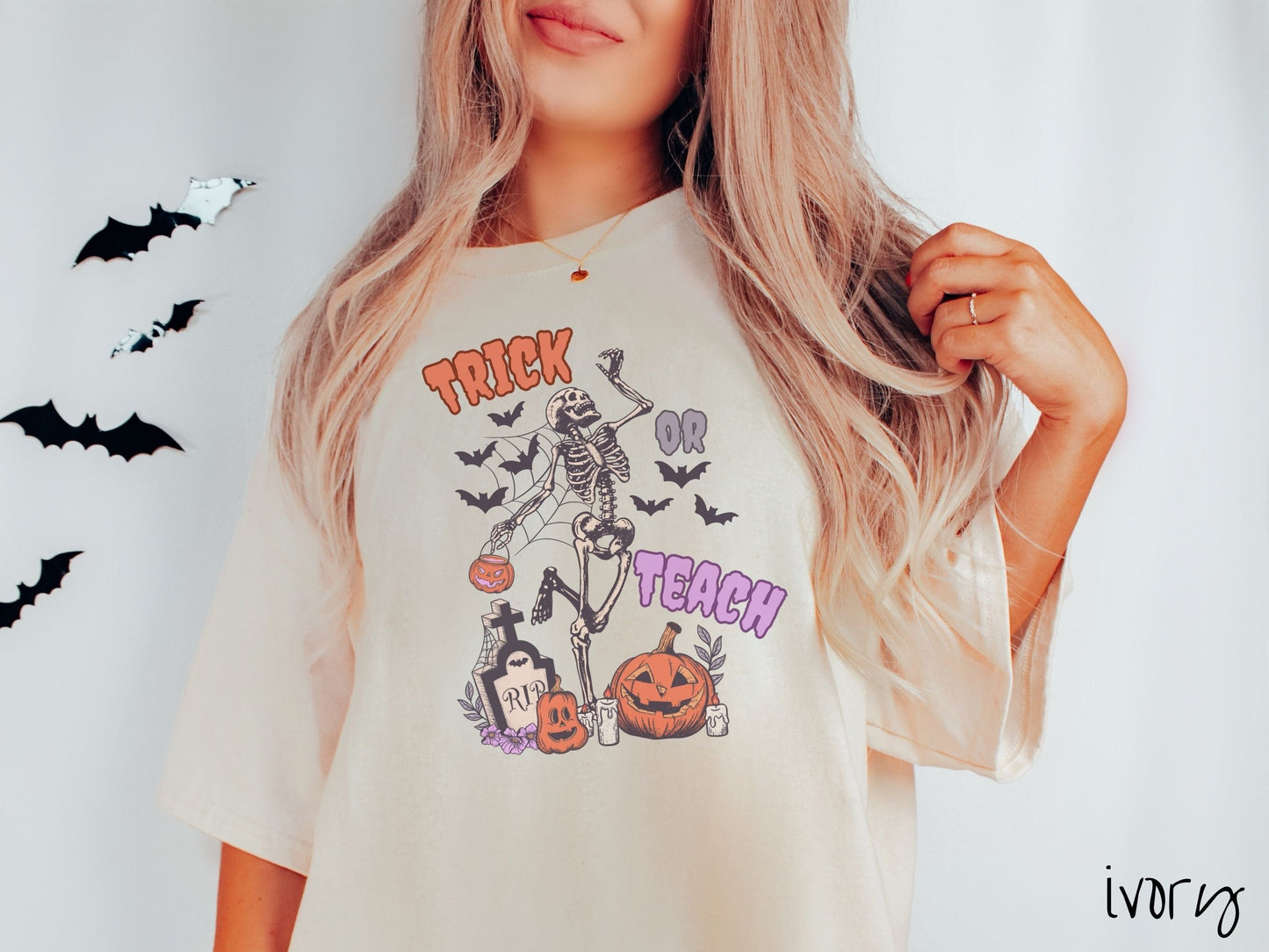 A woman wearing a cute, vintage ivory colored Comfort Colors T-shirt with text Trick or Teach in purple and orange font. There is a skeleton holding a pumpkin candy bucket dancing among spider webs and black bats over a tombstone and pumpkins.