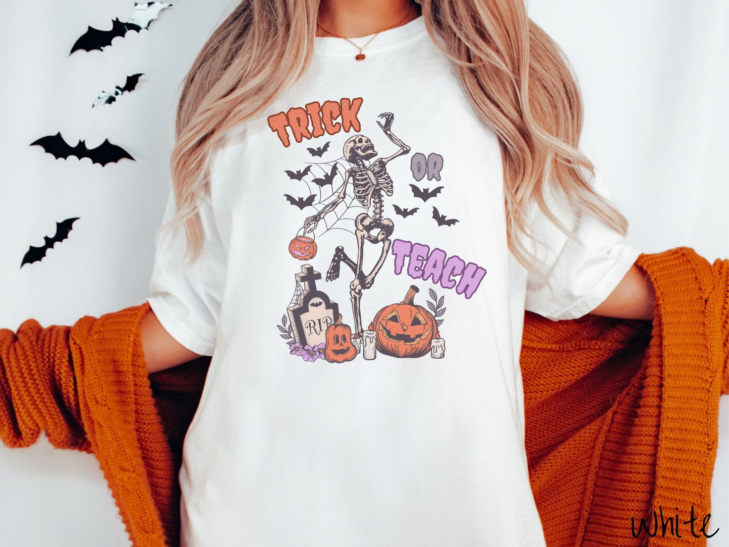 A woman wearing a cute, vintage white colored Comfort Colors T-shirt with text Trick or Teach in purple and orange font. There is a skeleton holding a pumpkin candy bucket dancing among spider webs and black bats over a tombstone and pumpkins.