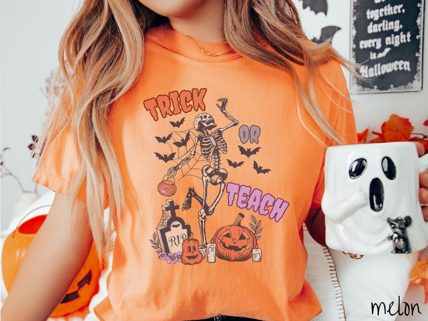 A woman wearing a cute, vintage melon colored Comfort Colors T-shirt with text Trick or Teach in purple and orange font. There is a skeleton holding a pumpkin candy bucket dancing among spider webs and black bats over a tombstone and pumpkins.