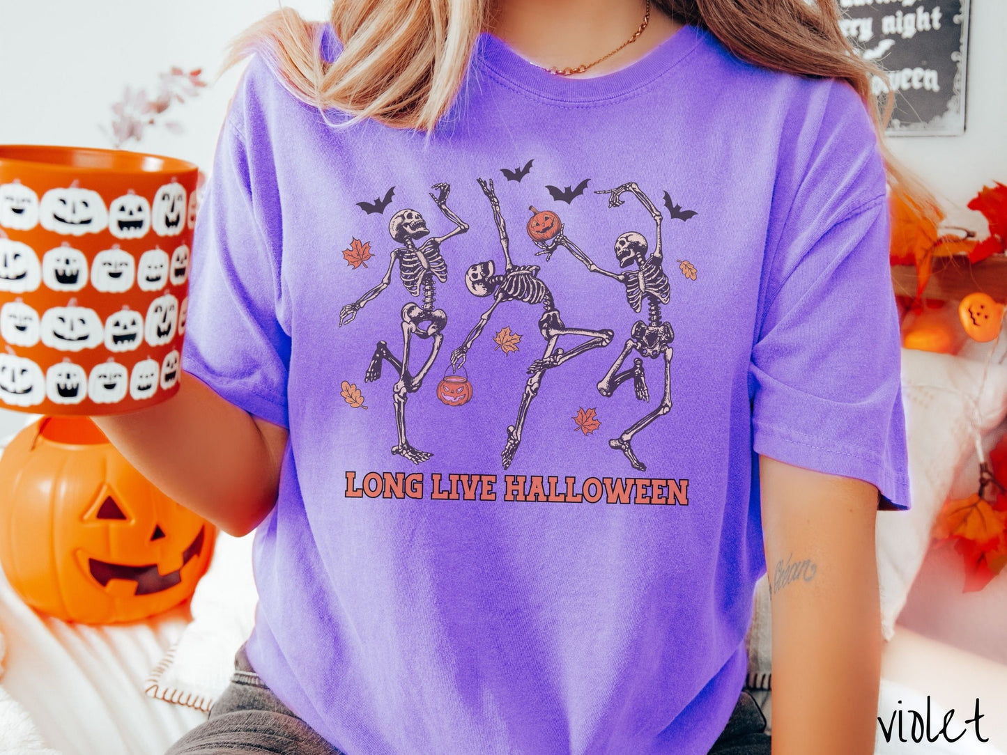A woman wearing a cute, vintage violet colored Comfort Colors T-shirt with the text Long Live Halloween in orange font. Above are three dancing skeletons holding pumpkins and jackolantern trick or treat buckets with bats and leaves in the air.