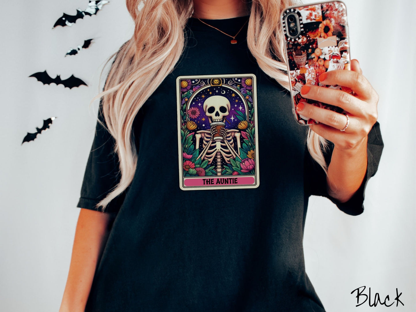 A woman wearing a cute, vintage black colored Comfort Colors T-shirt with text The Auntie along the bottom of a tarot card. Above is a smiling skeleton holding a to-go cup of coffee up to her mouth, and is surrounded by stars and colorful flowers.
