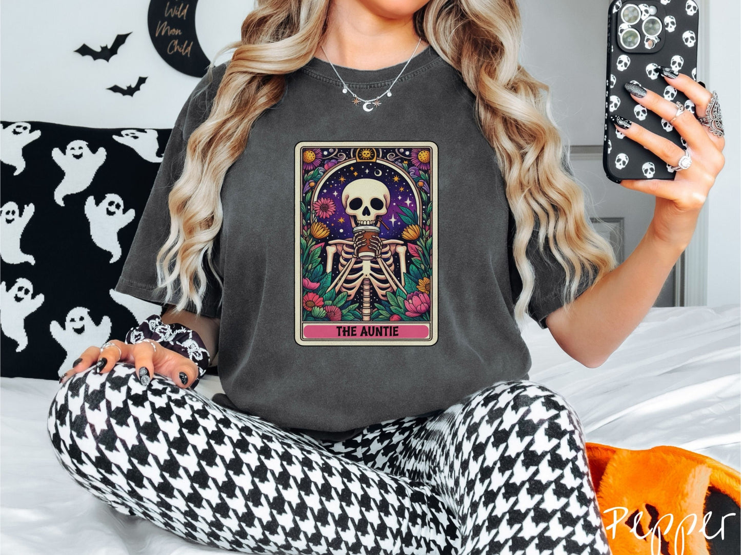 A woman wearing a cute, vintage pepper colored Comfort Colors T-shirt with text The Auntie along the bottom of a tarot card. Above is a smiling skeleton holding a to-go cup of coffee up to her mouth, and is surrounded by stars and colorful flowers.