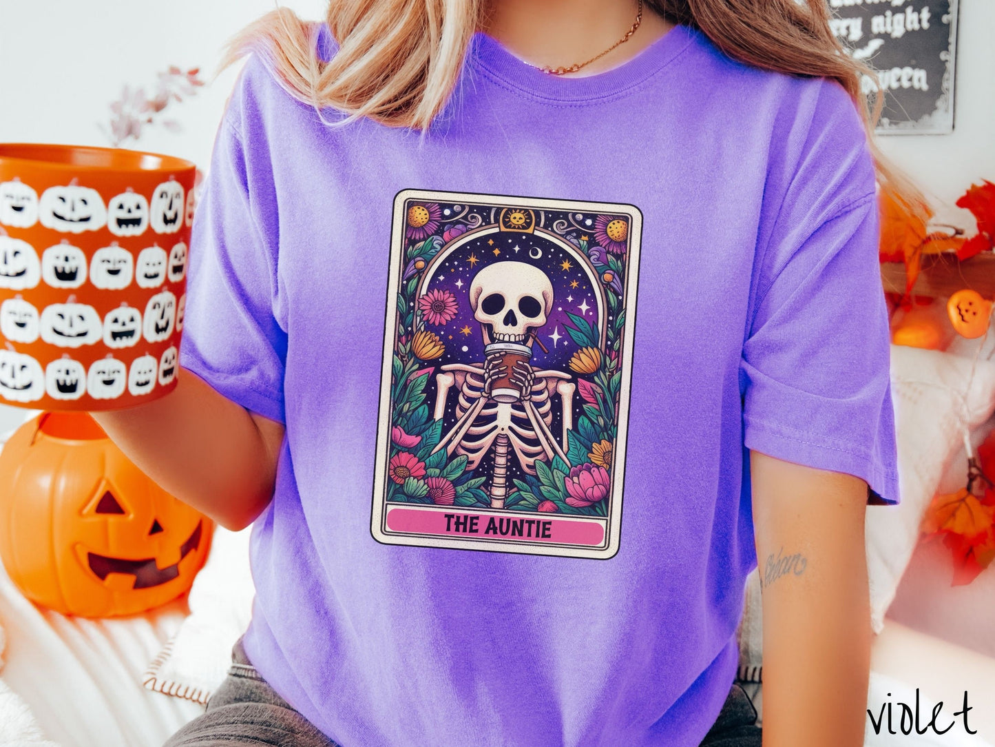A woman wearing a cute, vintage violet colored Comfort Colors T-shirt with text The Auntie along the bottom of a tarot card. Above is a smiling skeleton holding a to-go cup of coffee up to her mouth, and is surrounded by stars and colorful flowers.