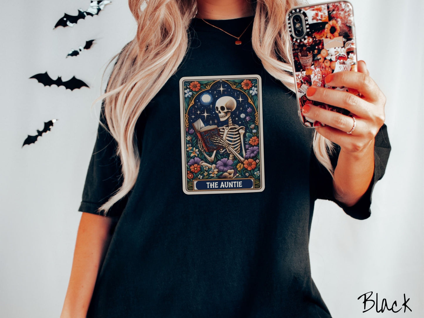 A woman wearing a cute, vintage black colored Comfort Colors T-shirt with text The Auntie along the bottom of a tarot card. Above is a skeleton reading an old looking book while under the moon and stars and surrounded by colorful flowers.