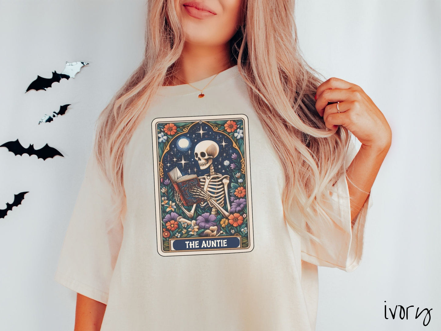 A woman wearing a cute, vintage ivory colored Comfort Colors T-shirt with text The Auntie along the bottom of a tarot card. Above is a skeleton reading an old looking book while under the moon and stars and surrounded by colorful flowers.