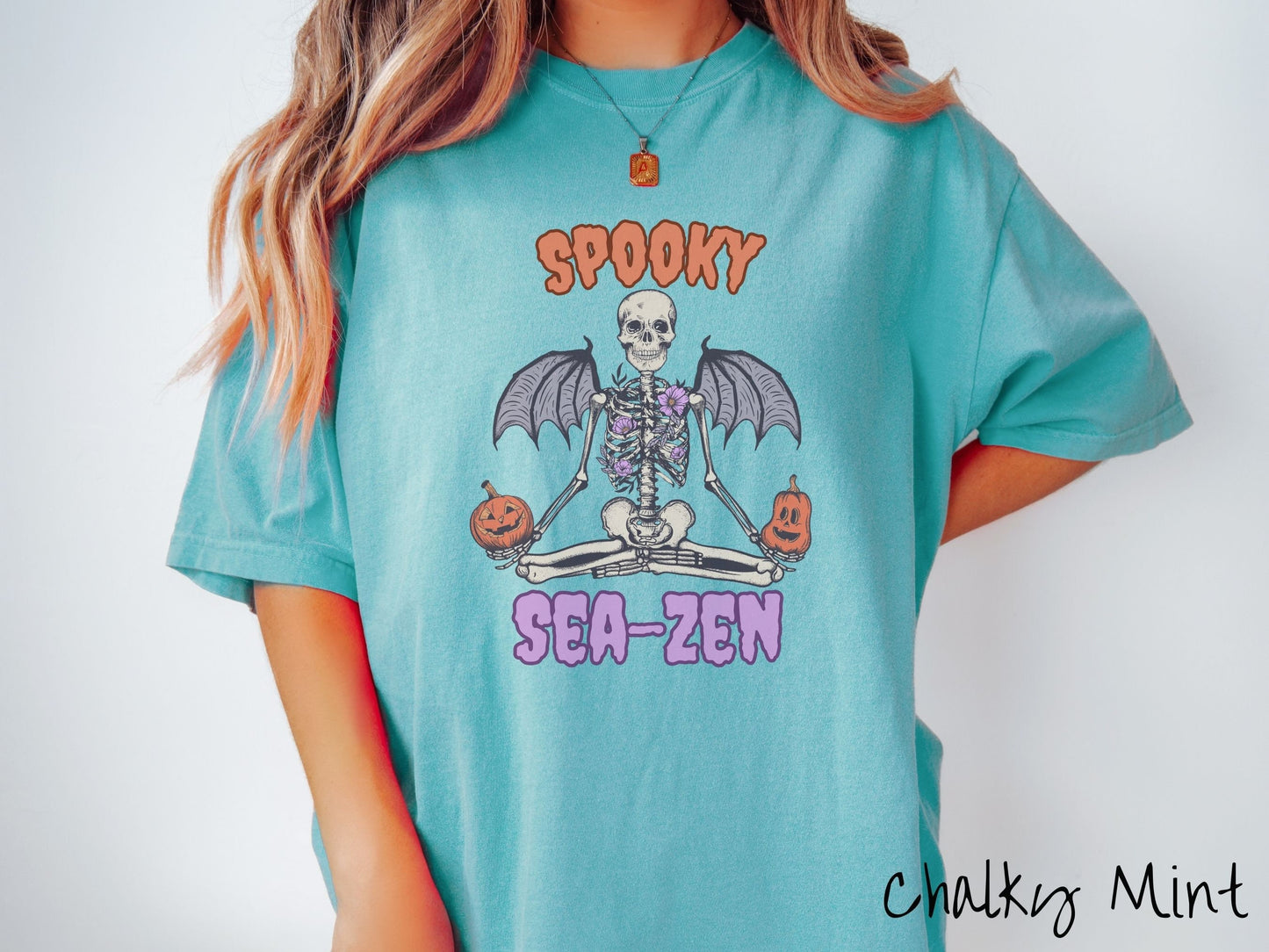 A woman wearing a cute, vintage chalky mint colored Comfort Colors T-shirt with text Spooky Sea-Zen in orange and purple font. There is a skeleton wearing flowers and bat wings sitting in a full lotus yoga pose while holding pumpkins in both hands.