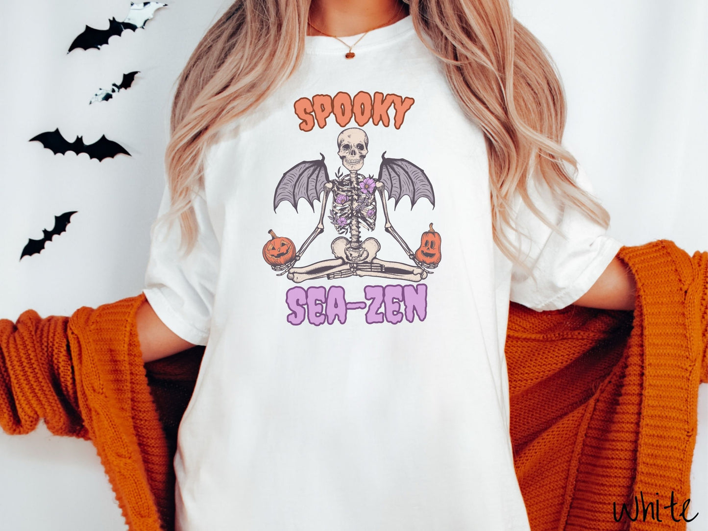 A woman wearing a cute, vintage white colored Comfort Colors T-shirt with text Spooky Sea-Zen in orange and purple font. There is a skeleton wearing flowers and bat wings sitting in a full lotus yoga pose while holding pumpkins in both hands.