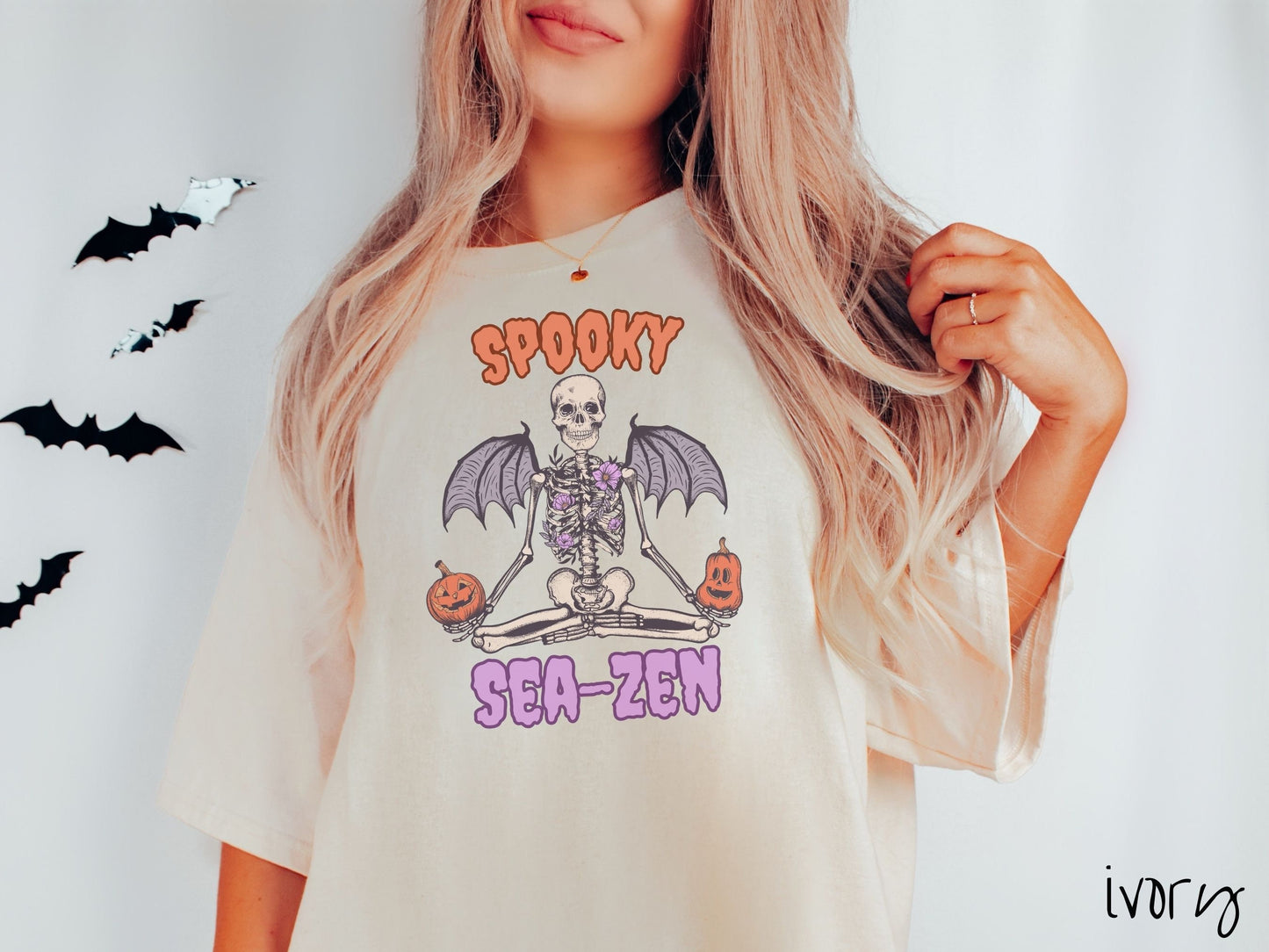 A woman wearing a cute, vintage ivory colored Comfort Colors T-shirt with text Spooky Sea-Zen in orange and purple font. There is a skeleton wearing flowers and bat wings sitting in a full lotus yoga pose while holding pumpkins in both hands.