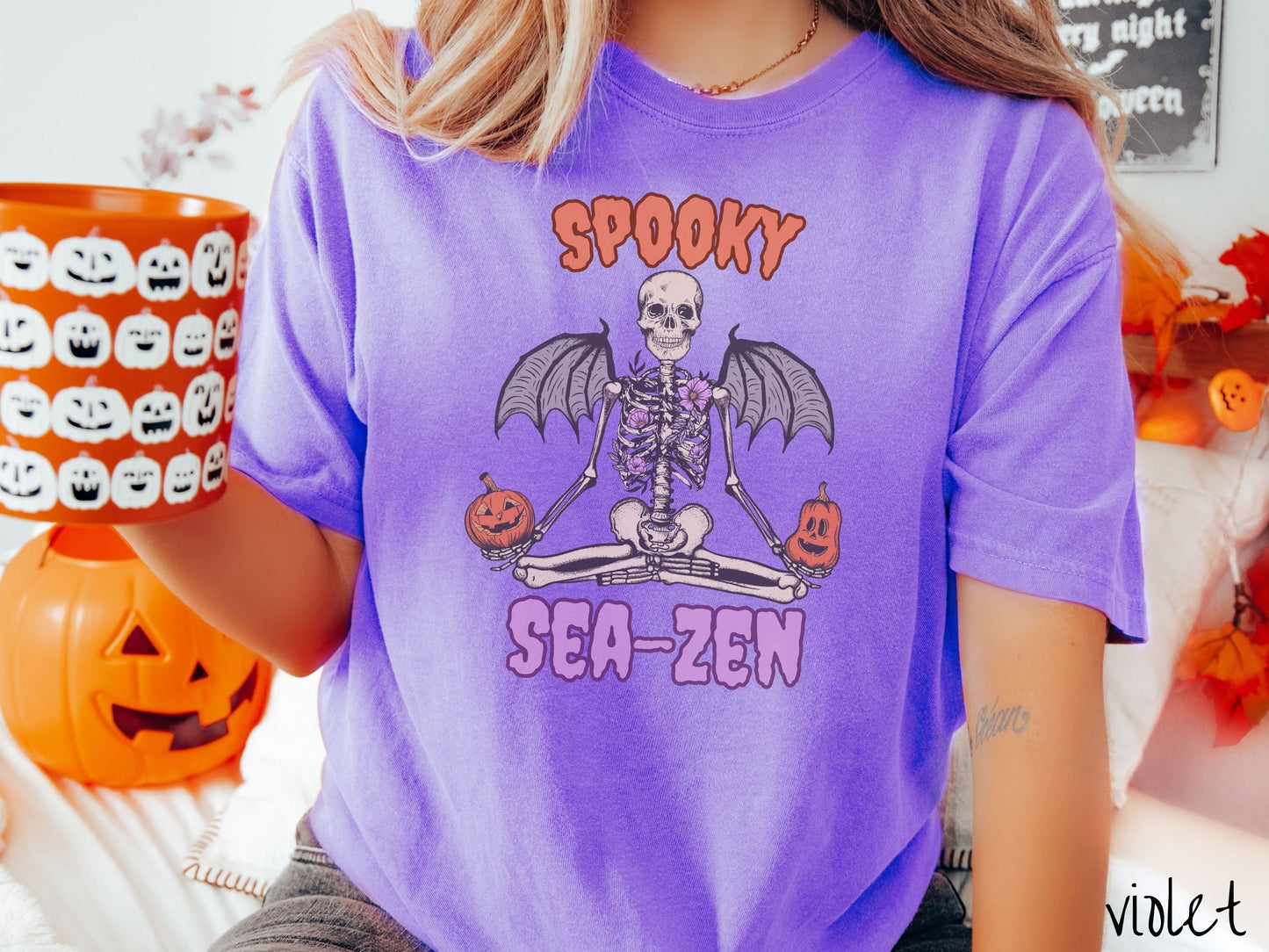 A woman wearing a cute, vintage violet colored Comfort Colors T-shirt with text Spooky Sea-Zen in orange and purple font. There is a skeleton wearing flowers and bat wings sitting in a full lotus yoga pose while holding pumpkins in both hands.