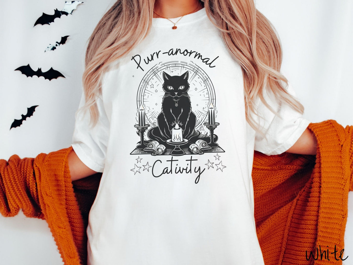 A woman wearing a cute, vintage white colored Comfort Colors T-shirt with text Purr-anormal Cativity in black font. There is a black cat sitting on an altar with candles and scrolls, staring straight forward with piercing eyes and wearing a necklace.
