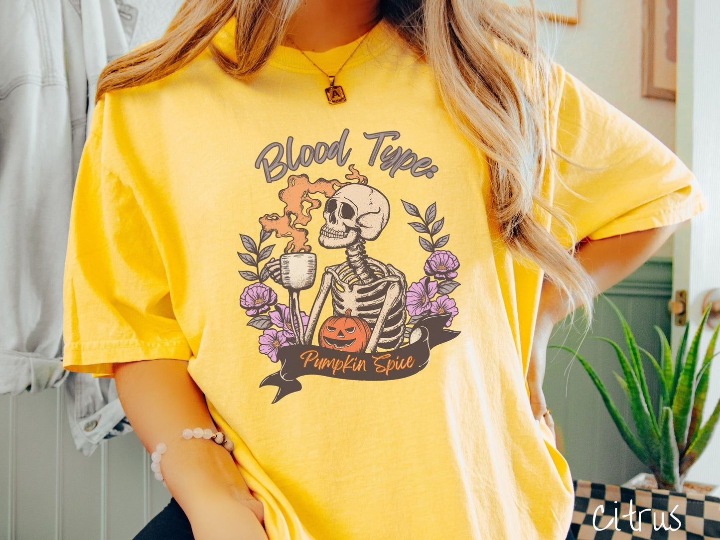 A woman wearing a cute, vintage citrus colored Comfort Colors T-shirt with text Blood Type: Pumpkin Spice. There is a skeleton holding a pumpkin spiced latte with orange steam coming out and a pumpkin while surrounded by purple flowers.