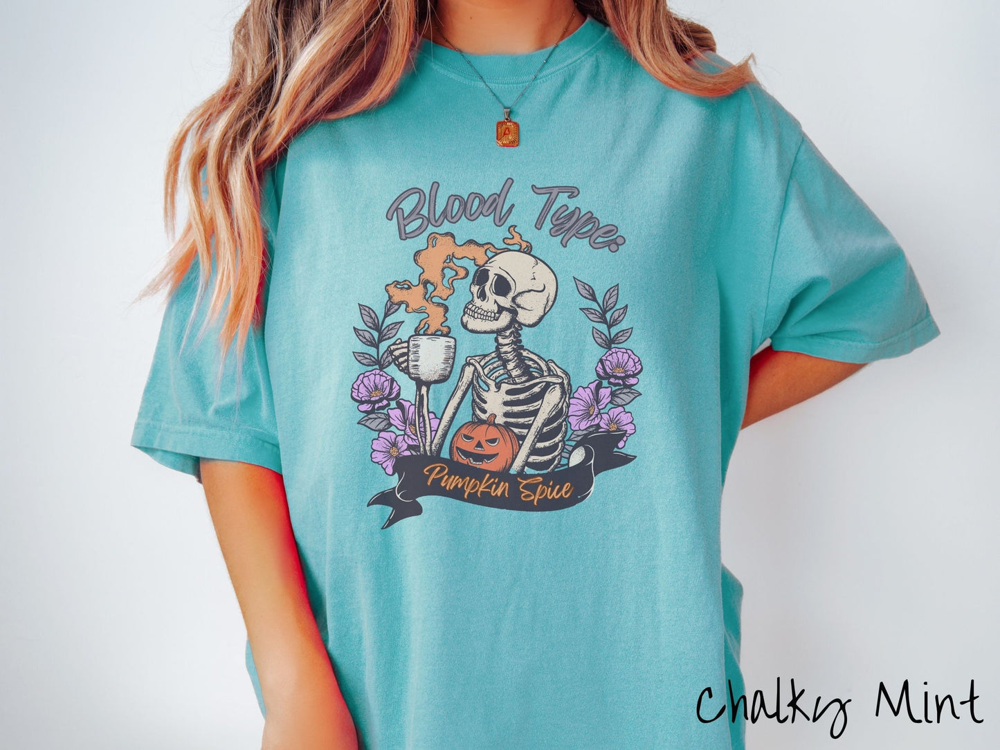 A woman wearing a cute, vintage chalky mint colored Comfort Colors T-shirt with text Blood Type: Pumpkin Spice. There is a skeleton holding a pumpkin spiced latte with orange steam coming out and a pumpkin while surrounded by purple flowers.