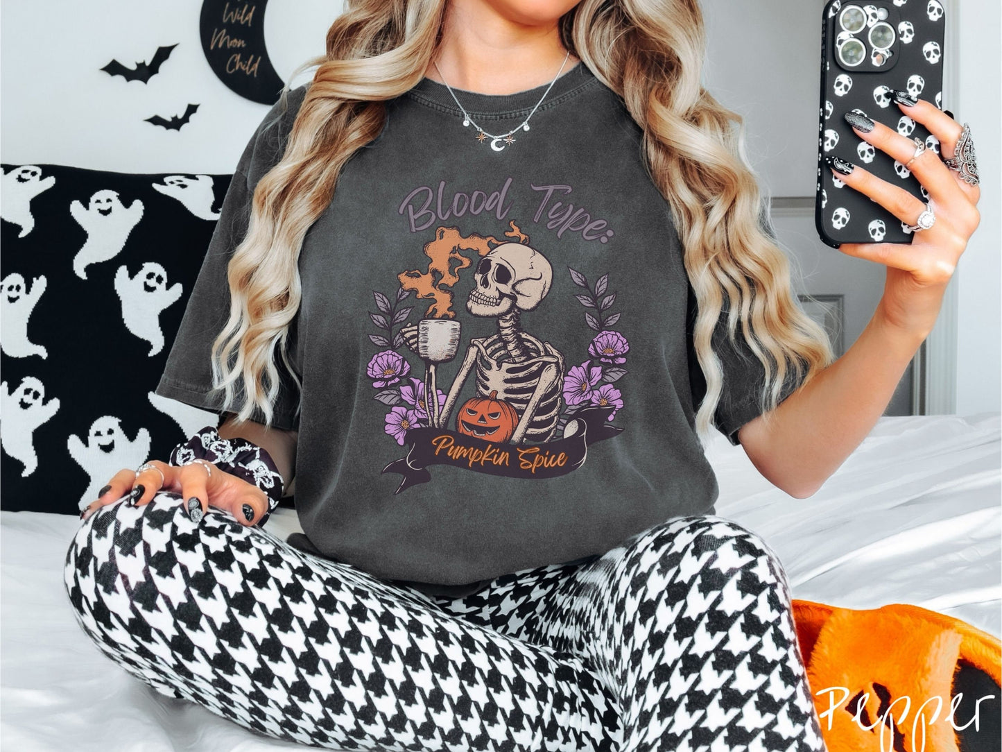 A woman wearing a cute, vintage pepper colored Comfort Colors T-shirt with text Blood Type: Pumpkin Spice. There is a skeleton holding a pumpkin spiced latte with orange steam coming out and a pumpkin while surrounded by purple flowers.