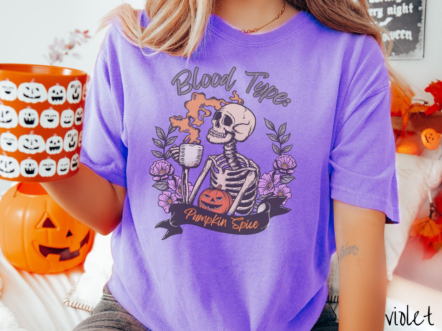 A woman wearing a cute, vintage violet colored Comfort Colors T-shirt with text Blood Type: Pumpkin Spice. There is a skeleton holding a pumpkin spiced latte with orange steam coming out and a pumpkin while surrounded by purple flowers.