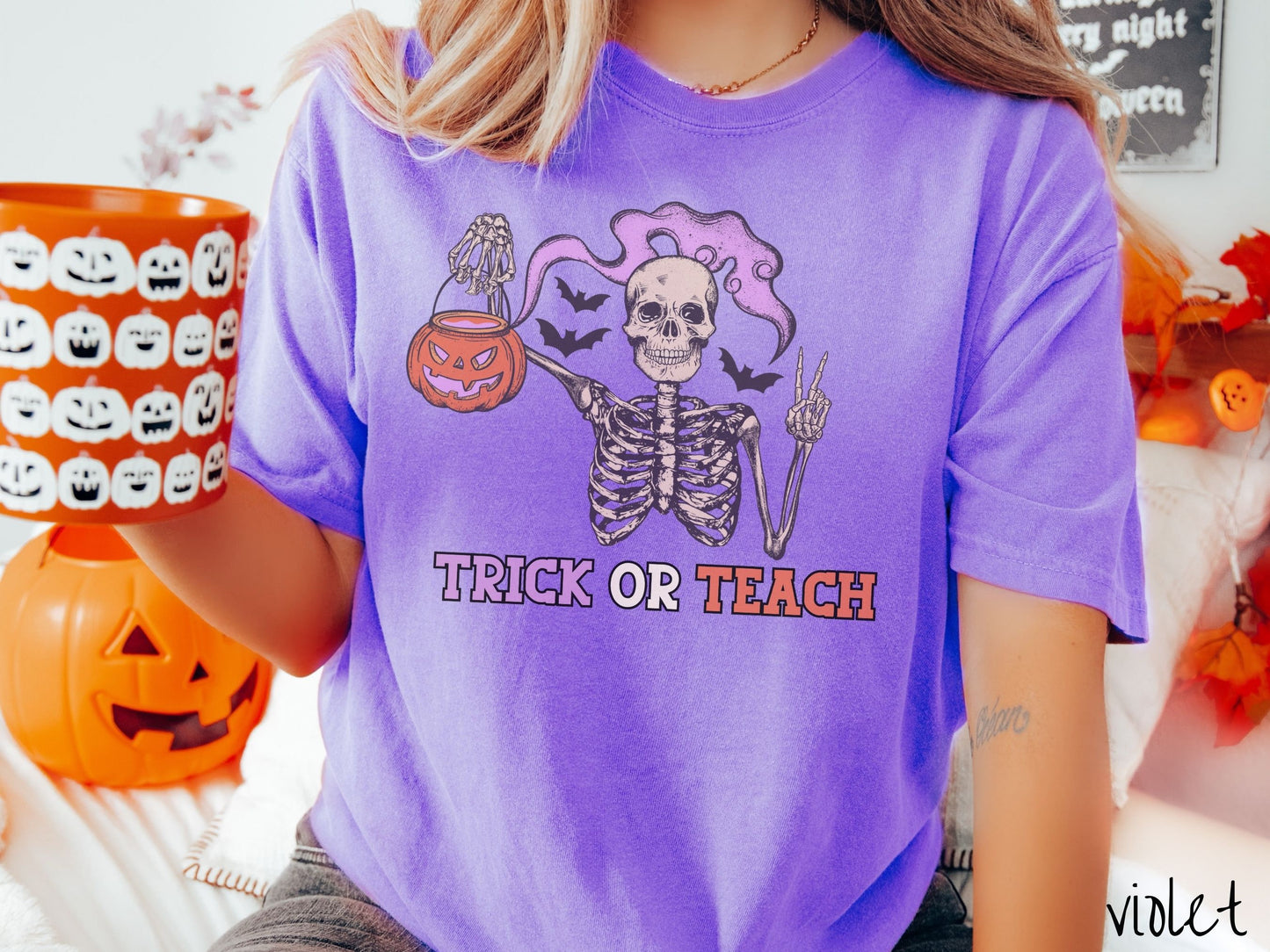 A woman wearing a cute, vintage violet colored Comfort Colors T-shirt with text Trick or Teach in purple and orange font. Above is a skeleton holding up a peace sign and a pumpkin candy bucket with purple smoke coming out and bats in the background.