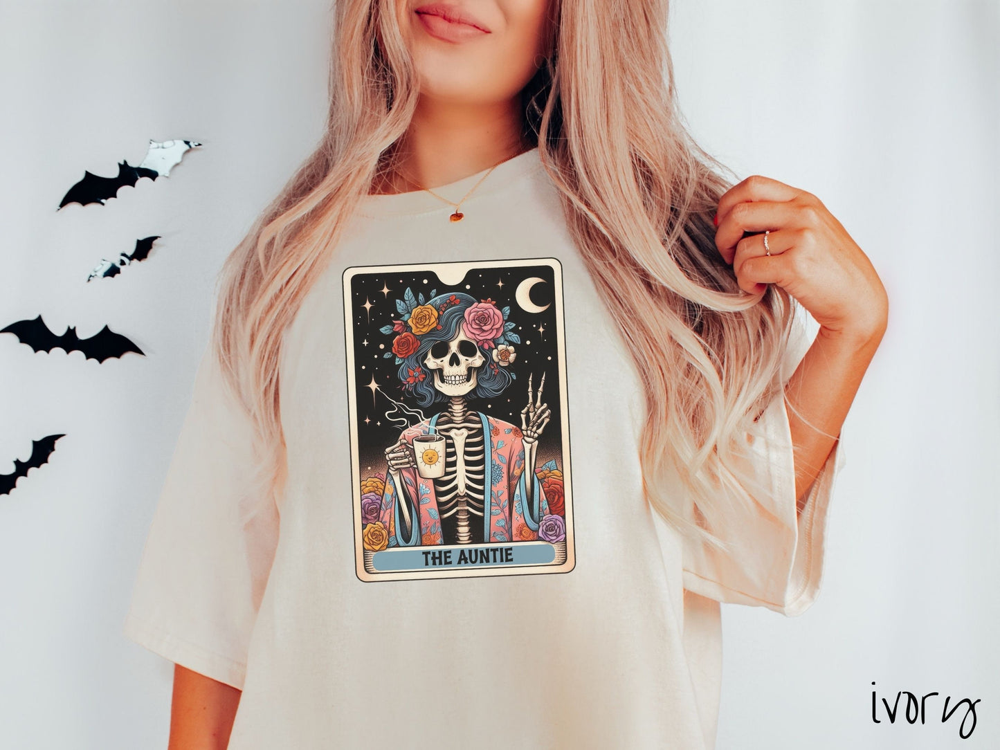 A woman wearing a cute, vintage ivory colored Comfort Colors T-shirt with text The Auntie along the bottom of a tarot card. Above is a picture of a female skeleton holding up a peace sign and wearing a flower robe holding a steaming cup of coffee.