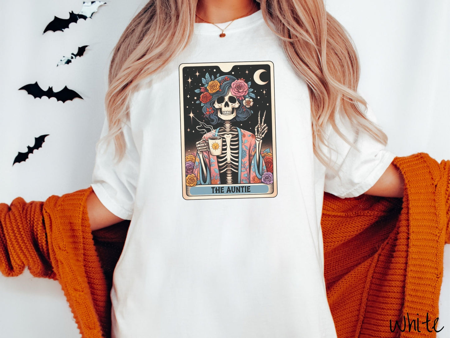 A woman wearing a cute, vintage white colored Comfort Colors T-shirt with text The Auntie along the bottom of a tarot card. Above is a picture of a female skeleton holding up a peace sign and wearing a flower robe holding a steaming cup of coffee.