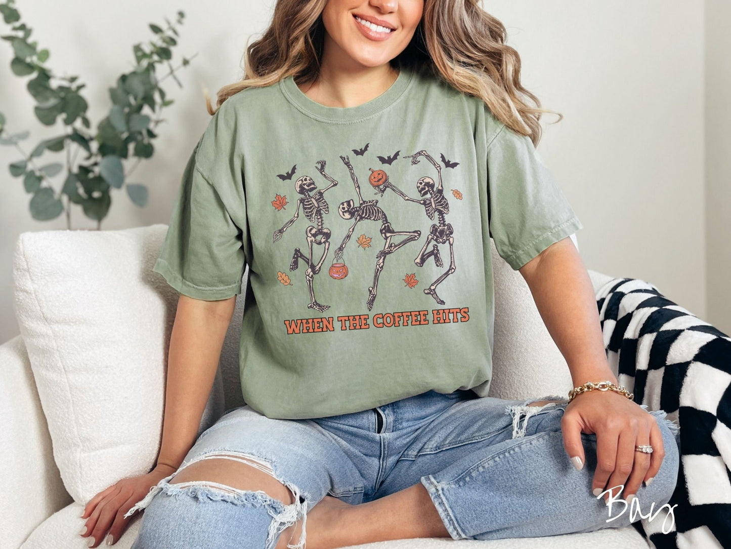 A woman wearing a cute, vintage bay colored Comfort Colors T-shirt with the text When the Coffee Hits in orange font. Above are three dancing skeletons holding pumpkins and jackolantern trick or treat buckets with bats and leaves in the air.