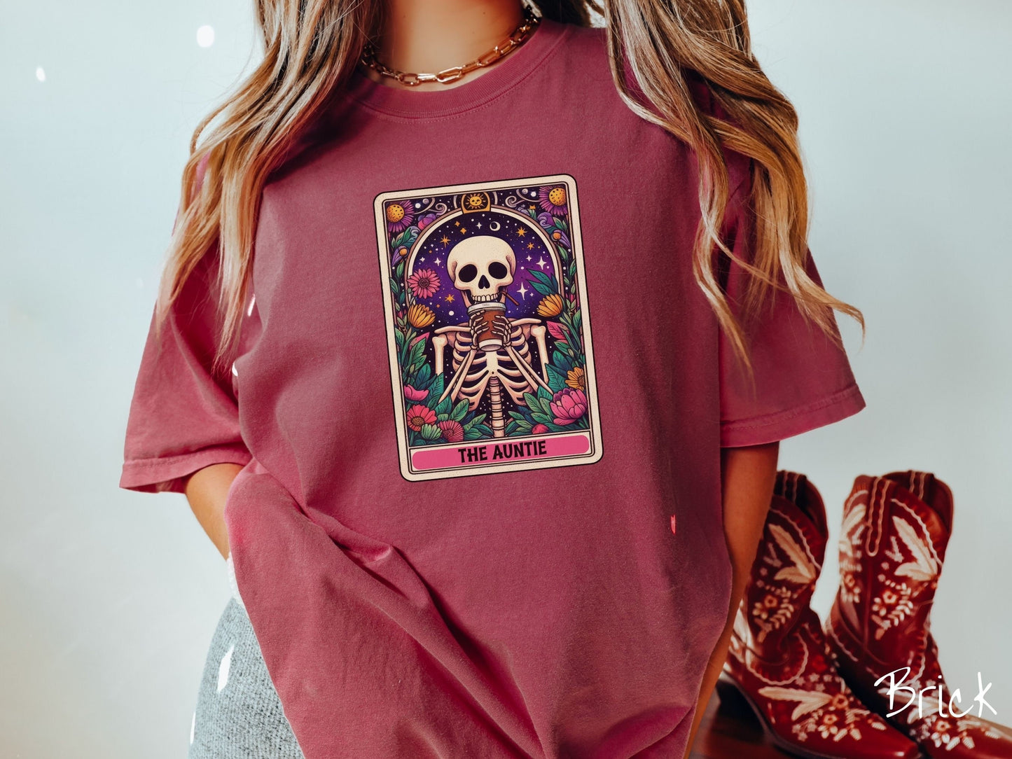 A woman wearing a cute, vintage brick colored Comfort Colors T-shirt with text The Auntie along the bottom of a tarot card. Above is a smiling skeleton holding a to-go cup of coffee up to her mouth, and is surrounded by stars and colorful flowers.