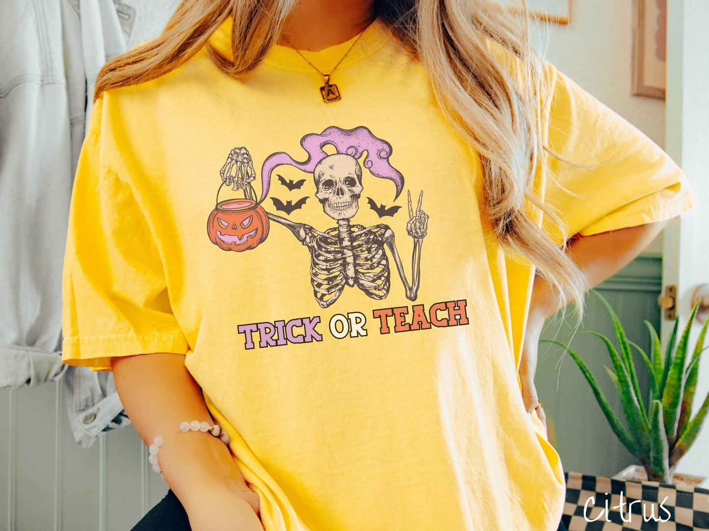 A woman wearing a cute, vintage citrus colored Comfort Colors T-shirt with text Trick or Teach in purple and orange font. Above is a skeleton holding up a peace sign and a pumpkin candy bucket with purple smoke coming out and bats in the background.